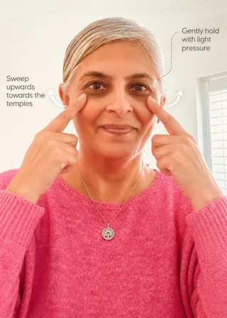 Neena demonstrating a face yoga exercise, gently pressing fingers to her temples